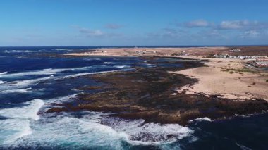 Fuerteventura kıyılarının, deniz fenerinin, tatil yerlerinin, gelgit havuzlarının ve sığ bir resifte kırılan dalgaların havadan görüntüsü. Lanzarote silueti ufukta beliriyor.