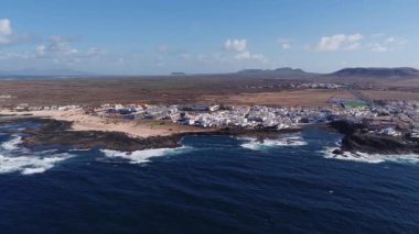 Fuerteventura 'da, kumlu sahil ile siyah volkanik sahil arasında, Atlantik dalgaları, gün ortası ışığı ve iç kısımlarda kurak tepeler bulunan, bembeyaz bir köyün hava görüntüsü..