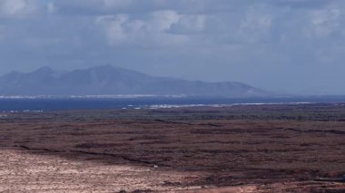 Fuerteventura kum tepelerinin, karanlık lav tarlalarının, kıvrımlı bir yolun, sahil sahanlığının, beyaz sörfün ve yumuşak bulut ışığıyla Lanzarote siluetinin hava günışığı görüntüsü..
