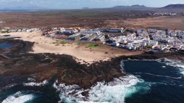 Siyah lav kayaları, soluk kumlar, beyazlatılmış evler, burunlarda sörf, gelgit havuzları, kum tepeleri ve uzak Lanzarote ile Fuerteventura kıyılarının hava manzarası..