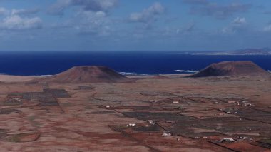 Fuerteventura üzerindeki hava 4K taramasında tephra konileri, pas ovaları, taş tarlaları, beyaz çiftlik evleri, toprak yolları, sörf, uzak Lanzarote, dağlar ve bulutlar görülüyor..