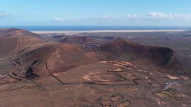 Hava taramasında volkanik koniler, krater jantları, lav duvarları, kum tepeleri, bir kasaba, park edilmiş arabalar, toprak yol, mavi okyanus ve ufukta Lanzarote görülüyor.
