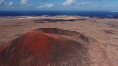 Hava aracı Fuerteventura 'daki kırmızı bir volkanik kraterin üzerinde süzülüyor, dik yamaçlar ve dairesel oyuklar kurak bir ovaya hükmediyor, kıyı kasabası ve Lanzarote denizin ötesinde beliriyor..