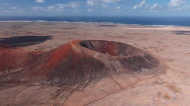 Fuerteventura 'da siyah lav çizgileri olan paslı kırmızı bir kraterin üzerini hava temizliyor. Toprak izleri, taş duvarlar, küçük koniler ve sahil yerleşimi yumuşak bulutların altında belirir..