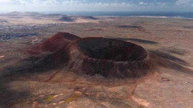 Hava görüntüsü Fuerteventura 'da bir kase şeklinde volkanik krater, kırmızı ve siyah akrep yamaçları, ikincil bir koni, taş duvarlar, muhafazalar, bir kıyı köyü ve Atlantik.