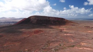 Hava 4K taraması Kuzey Fuerteventura 'da kırmızı ve siyah bir volkanik krater gösteriyor. Küçük bir kıyı kasabası, Atlantik ufku, aşınmış çember ve taş duvarları var..