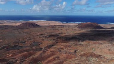 Hava 4K görüntüsü Fuerteventura 'da iki kül konisi, pas kırmızısı lav ovası, kumlu sahil ve Atlantik sörfü gösteriyor..
