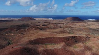 Hava aracı Fuerteventura, Kanarya Adaları 'ndaki pas kırmızısı bazaltik ovanın üzerinde süzülerek küçük kraterler, iki düz koni, sörf ve gün ortası bulutları ortaya çıkarıyor..