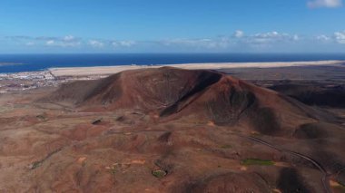Hava kayması, paslı kırmızı bir volkanik krateri, lav tarlalarını ve toprak bir yolu ortaya çıkarır. Fuerteventura 'daki Corralejo kum tepeleri ve köy Atlantik' le buluşuyor, Lanzarote ufukta..