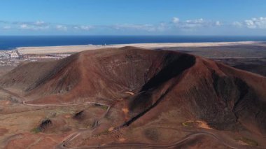 Havadan bakıldığında kırmızı kahverengi bir krater, lav tarlaları ve Corralejo kumullarına ve kıyı kasabasına giden izler görülüyor..