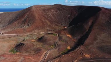 Hava görüntüsü Fuerteventura üzerinde lav çizgileri ve kül konileri olan paslı kırmızı bir krater gösteriyor. Kirli bir yol ve park edilmiş 4x4 'ler içeride, ufukta Atlantik görünüyor..