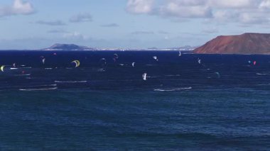 Atlantik 'in kuzey Fuerteventura açıklarında gündüz görüşü, ufukta Lanzarote, kırmızı volkanik koni, düzinelerce uçurtma ve binici uyanma çizgileri ve sprey ile kavisler oyar.
