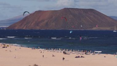Fuerteventura 'daki geniş kumlu sahil manzarası, uçurtmalar ve uçurtmacılar derin mavi dalgalar, pas rengi volkanik koniler, ufukta Lanzarote, yelkenliler, şezlonglar, şemsiyeler.