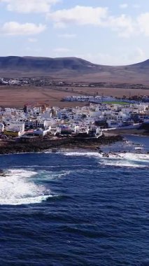 Atlantik dalgaları siyah volkanik kayalara, düşük mesafelere, tepeler gibi kratere ve yavaş hareket eden volkanik kayalara çarptığında Fuerteventura 'daki bembeyaz bir sahil kasabasının gündüz görüntüsü.