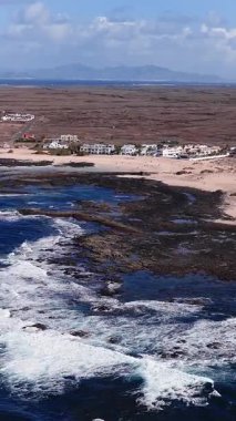 Fuerteventura kıyılarının havadan görünüşü, Atlantik dalgaları siyah lavlar üzerinde, soluk kumlu plajlar, beyaz tatil evleri, gelgit havuzları, uzak koniler ve Lanzarote görünür.