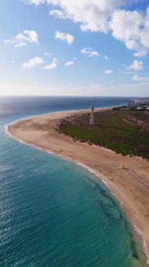 Morro Jable deniz feneri, kumlu tükürük, tatil köyleri ve Fuerteventura 'da rüzgar türbinleri olan kahverengi tepeler. Turkuaz Atlantik turları güneşli plajlarda güneşleniyor..