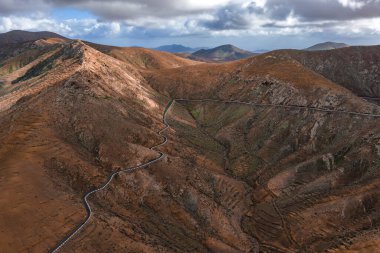 İspanya, Fuerteventura 'da, Caldera Los Arrabales yakınlarında, kırsal tepeler ve kırgın bulutların altında serin bir öğleden sonra ışığı altında uzanan dolambaçlı bir dağ yolu manzarası..