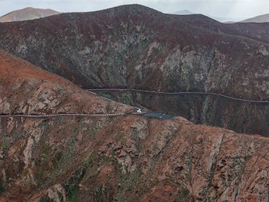 Fuerteventura, Kanarya Adaları 'ndaki dar bir dağ geçidinin hava manzarası, yılansı bir yol, katılımda park edilmiş arabalar, ve bulutlu ışık altında katmanlı lav duvarları.