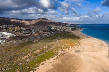 Hava sahnesi Fuerteventura, Kanarya Adaları 'nda Morro Jable' ı gösteriyor. Turkuaz su uzun kumlarla, tesis kompleksleriyle, volkanik tepelerle, gelgit ovalarıyla ve hareket eden bulut gölgeleriyle buluşur..