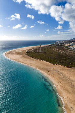 Hava sahnesinde Morro Jable 'ın Fuerteventura' daki plaj yayı, beyaz Morro Jable Deniz feneri, turkuaz Atlantik, şezlonglar, cankurtaran kulesi, tatil köyleri ve öğle vakti bulutları görülüyor..
