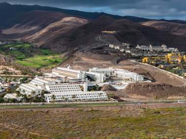 Hava görüntüsü, karanlık volkanik tepelerde beyaz teraslı oteller, canlı golf sahaları ve Fuerteventura 'da alçak bulutların altında arabaları ve otobüsleri olan bir kıyı otoyolu gösteriyor..