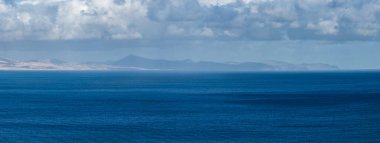 Derin kobalt okyanusu ufukta sisli Fuerteventura ve Lanzarote ile buluşur. Dalgalanan bulutlar ve yumuşak gün ışığı, uzun lens sıkıştırma ve minimalist kompozisyon.