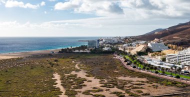 Teraslı beyaz oteller, Fuerteventura 'daki Morro Jable' da uzun ve kumlu bir sahil kenarında bir avuç içi geçidi hizasında. Volkanik kahverengi tepeler gün ortası ışığı ve dağınık bulutlar altında yükselir..