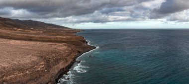 Fuerteventura 'nın batı kıyısındaki çorak uçurumlar Playa del Bajo de la Burra yakınlarında deniz mavisi sığ, derin mavi su ve kara kara düşünen bulutlarla Atlantik' le buluşur..