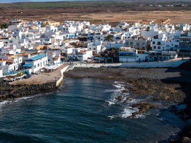 Fuerteventura 'daki kayalık bir körfezdeki beyaz düz çatı binalarının hava manzarası. Derin mavi Atlantik, siyah lav kayalarıyla, küçük rıhtımlarla ve parlak gün ışığı altında gezinmeyle karşılaşıyor..