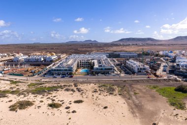 Havadan bakıldığında kumlu kumsalda havuzları, vinçleri ve inşaatı, karanlık volkanik ovaları, tepeler, köyler gibi kalderaları ve açık gün ortası gökyüzü olan beyaz, alçak daireler görülüyor..