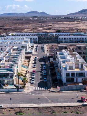 Fuerteventura 'daki beyaz kübik ev ve park halindeki arabaların hava manzarası, önümüzde karanlık bir cephe binası, ötelerinde volkanik koniler, gün ortası ışığı, sağda devam eden inşaat.