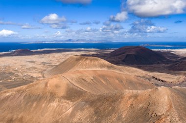 Hava sahnesinde Caldera Los Arrabales ve Playa del Bajo de la Burra yakınlarındaki Fuerteventura konileri ve kalderaları, Lanzarote ana hatları, sahil kasabası ve gün ortası altında tuzlu beyaz plajlar görülüyor..