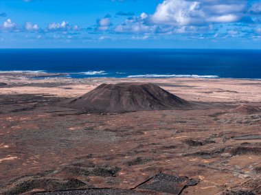 Fuerteventura, Kanarya Adaları 'ndaki, bazaltik tarlaları, bej kum tepeleri ve Atlantik Okyanusu' nun öğlen ışığı ve kümülüs bulutları altındaki tek bir kül konisinin havadan görüntüsü..