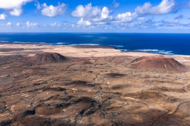 Havadan bakıldığında Caldera Los Arrabales yakınlarındaki bir lav ovasında iki kırmızı kahverengi koni görünüyor. Atlantik sörfü soluk kumlarla buluşuyor. Gün ortasında kümülüs bulutları yumuşak gölgeler bırakıyor..