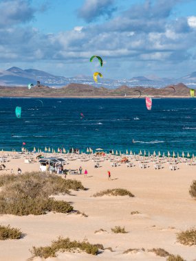 Solgun kum tepecikleri derin mavi sularda renkli uçurtmalarla karşılaşıyor. Uçurtma sörfçüleri kar dalgaları gibi. Şemsiye ve şezlonglar kıyı boyunca dizili. Lanzarote tepeleri karşıya oturur..
