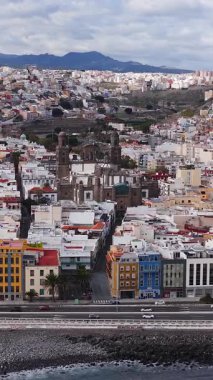 Las Palmas de Gran Canaria 'nın Katedral de Santa Ana ile gündüz görüntüsü, arabalar kıyı yolunda hareket ediyor, dalgalar kayalık bir kıyıya, bulutlu bir gökyüzüne ve uzak tepelere ulaşıyor