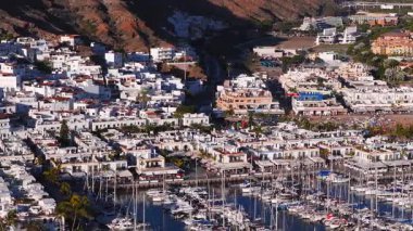 Hava 4K görüntüsü, öğleden sonra geç saatlerde Porto de Mogan, Gran Canaria 'da beyaz teraslı binalar, yoğun bir marina, kumlu plaj ve palmiye gezintisi gösteriyor..