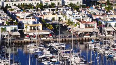 Beyaz evler, yeşil kepenkler, palmiye ağaçları, demirli yatlar, durgun sular ve gün batımında gezinti güvertesindeki insanlarla birlikte Puerto de Mogan, Gran Canaria 'nın hava 4K sahnesi..
