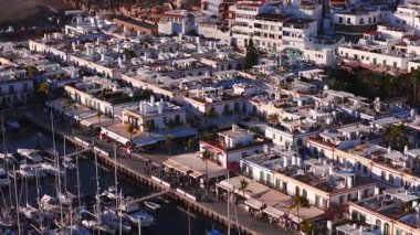 Büyük Kanarya 'daki Puerto de Mogan' ın 4K hava görüntüsü. Yelkenli liman, palmiyeli gezinti güvertesi, kafeler, market tezgahları ve öğleden sonra aydınlığında beyaz teraslı çatılar..
