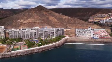Güney Gran Canaria tatil beldelerinin hava manzarası, Radisson Mavisi tabelası, kumlu bir körfez kenarındaki palmiyeler, havuzlar ve avralar, tekneler, deniz kenarı, kurak volkanik tepeler, parçalı bulutlu ışık tepeleri
