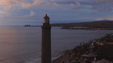 Hava aracı Faro de Maspalomas, Gran Canaria 'da sürüklenir. Palm Resort, gezinti ışıkları, tekneler, hafif dalgalar ve uzak Teide Dağı silueti alacakaranlıkta belirir..
