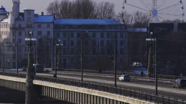 Riga, Letonya 'daki hava sahnesinde, güneşli bir kış gününde trafik, tramvaylar, yayalar ve dönme dolap bulunan tozlu bir köprü görülüyor..