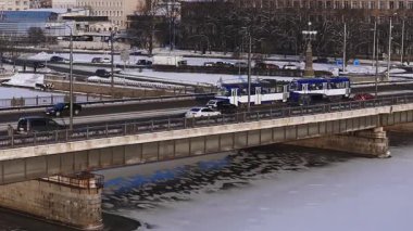 Riga, Letonya 'daki 4K hava sahasında Daugava Nehri, bir taş ve çelik yol ve tramvay köprüsü, mavi ve beyaz tramvaylar, sabit trafik ve tarihi kilise kuleleri görülüyor..