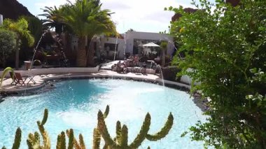 Ziyaretçiler, Kanarya Adaları 'ndaki Lagomar Müzesi' nde dairesel bir turkuaz havuz kenarında otururken, kavisli su jetleri parıldıyor, öğlen ışığı parlıyor ve kaktüs ve palmiye çerçeveli lav kayaları..