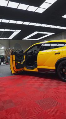 Kamera kırmızı kattan kabine doğru kayıyor, kapı açık, siyah alaşımlı tekerlek ve Performante pervazı görünür, LED paneller stüdyo garajındaki parlak sarı gövdeyi yansıtıyor..