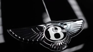 Stüdyoda krom kanatlı B kaput amblemi çerçeveyi doldurur. Softbox yansımaları siyah ayna boyası üzerinde süzülürken kamera yavaş yavaş açıları izler ve kayar.