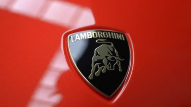 Lamborghini kalkanının metalik turuncu kaputtaki makro görüntüsü, yavaş tavalar ve ince raf odaklı bir stüdyoda, yumuşak kutu yansımaları boyada süzülüyor.