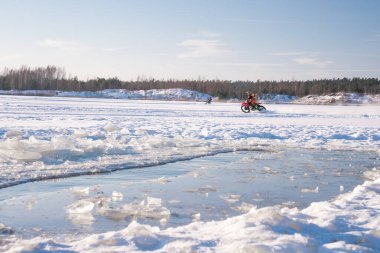 İki motosikletçi karla kaplı buzun üzerinde, buzlu buzun yanında kavisli kavisler ve açık bir kurşun, düşük güneş ışığı, açık gökyüzü ve orman kıyıları.