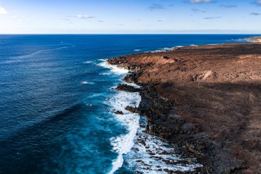 Hava sahnesinde Lanzarote, Kanarya Adaları, Atlantik dalgaları Timanfaya yakınlarındaki bazalt kayalıklara çarpıyor. Lav alanları, gün ortası ışığı altında iç kesimlere kadar uzanıyor..