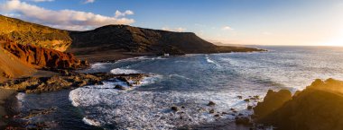 El Golfo, Lanzarote yakınlarındaki engebeli volkanik kayalıklar ve sörfler koyu lav kayaları, kırmızı tuff, siyah kum ve sıcak öğleden sonra ışığı altında köpüren dalgaları gösteriyor..